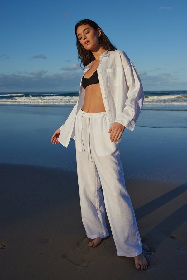 Woman on posing on the beach win white linen pants and shirt with a black bralette